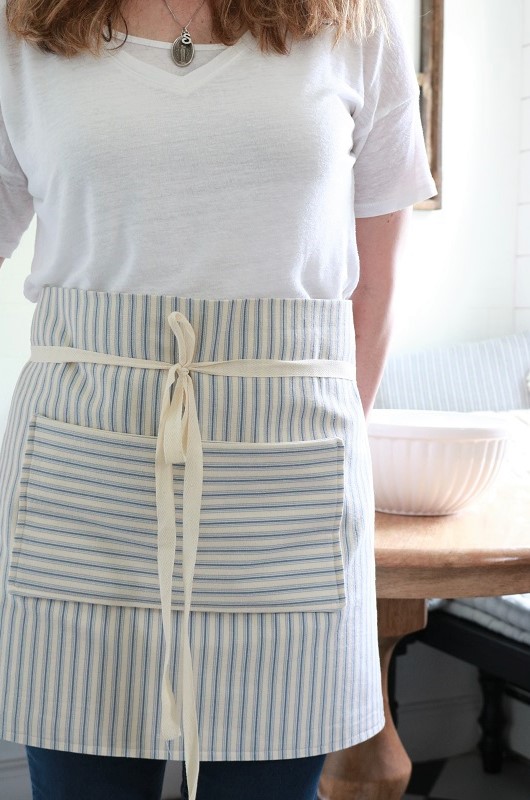 Farmhouse Style Pocket Apron - wearing in kitchen, at table, facing out with hand out of pocket